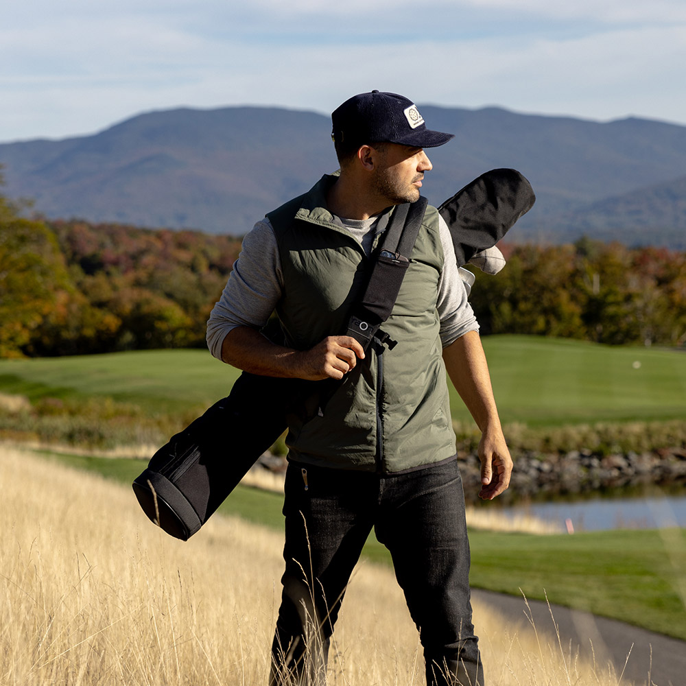An athletic man dressed in casual street attire stands on a grassy hill in a beautiful golf course with the Sunday Sling golf bag strapped to his back
