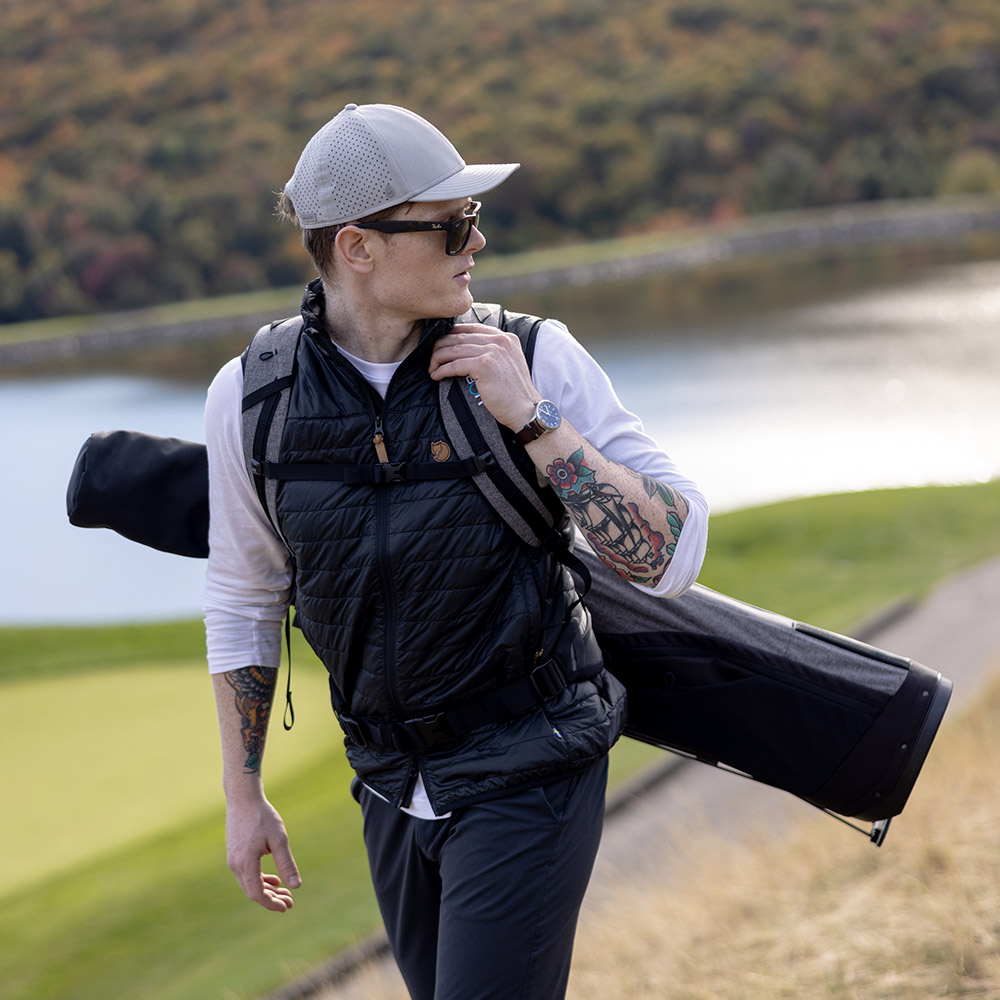 A young man wearing trendy clothing and having tattoos on his arm carries his golf clubs in the Golf Pack as he walks the golf course